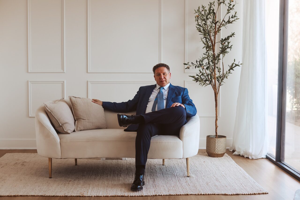 Attorney Carlos Maury in a navy suit, seated on a cream sofa beside an olive tree.
