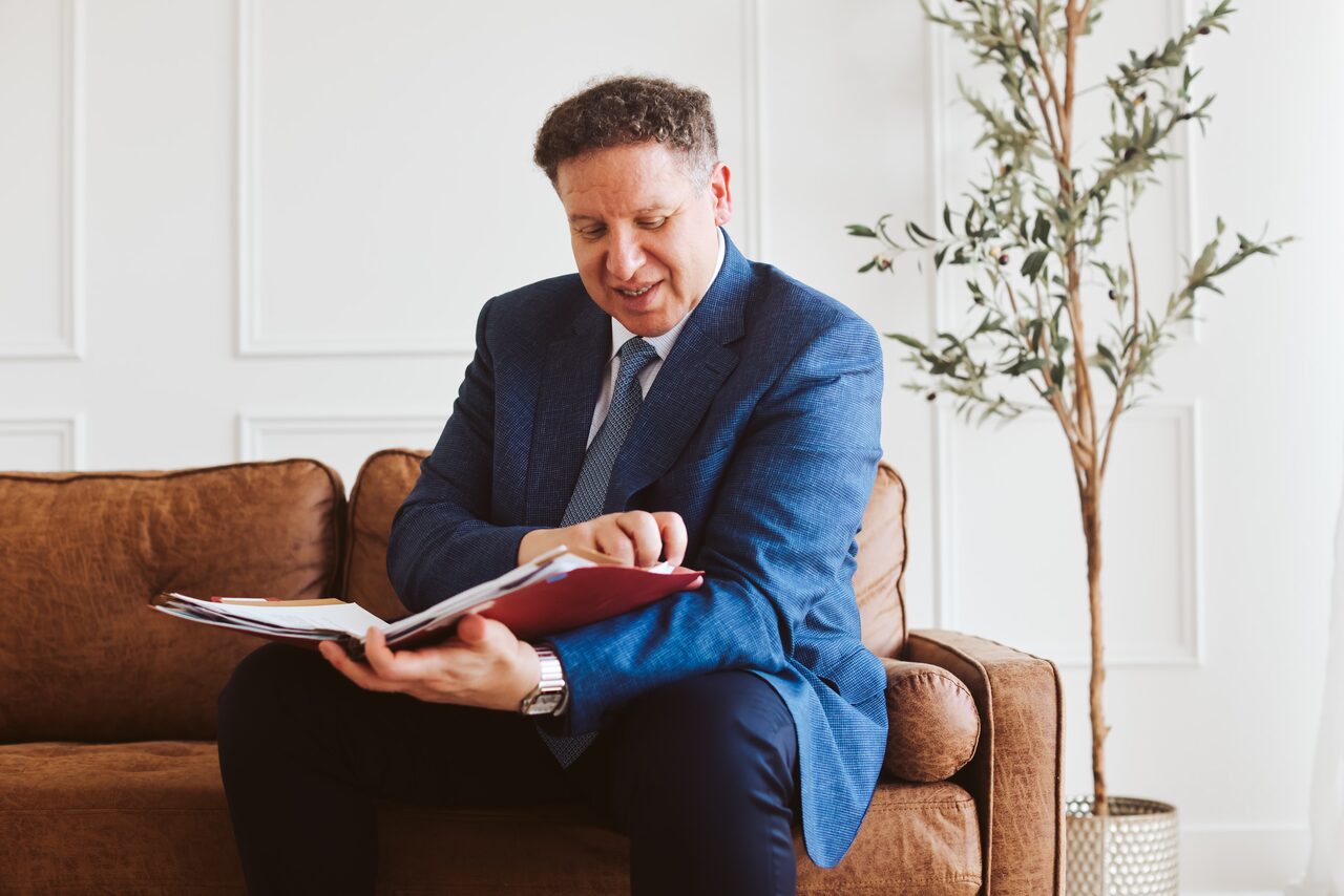 Attorney Carlos Maury, in a blue suit, reviewing a case file while seated on a leather sofa.